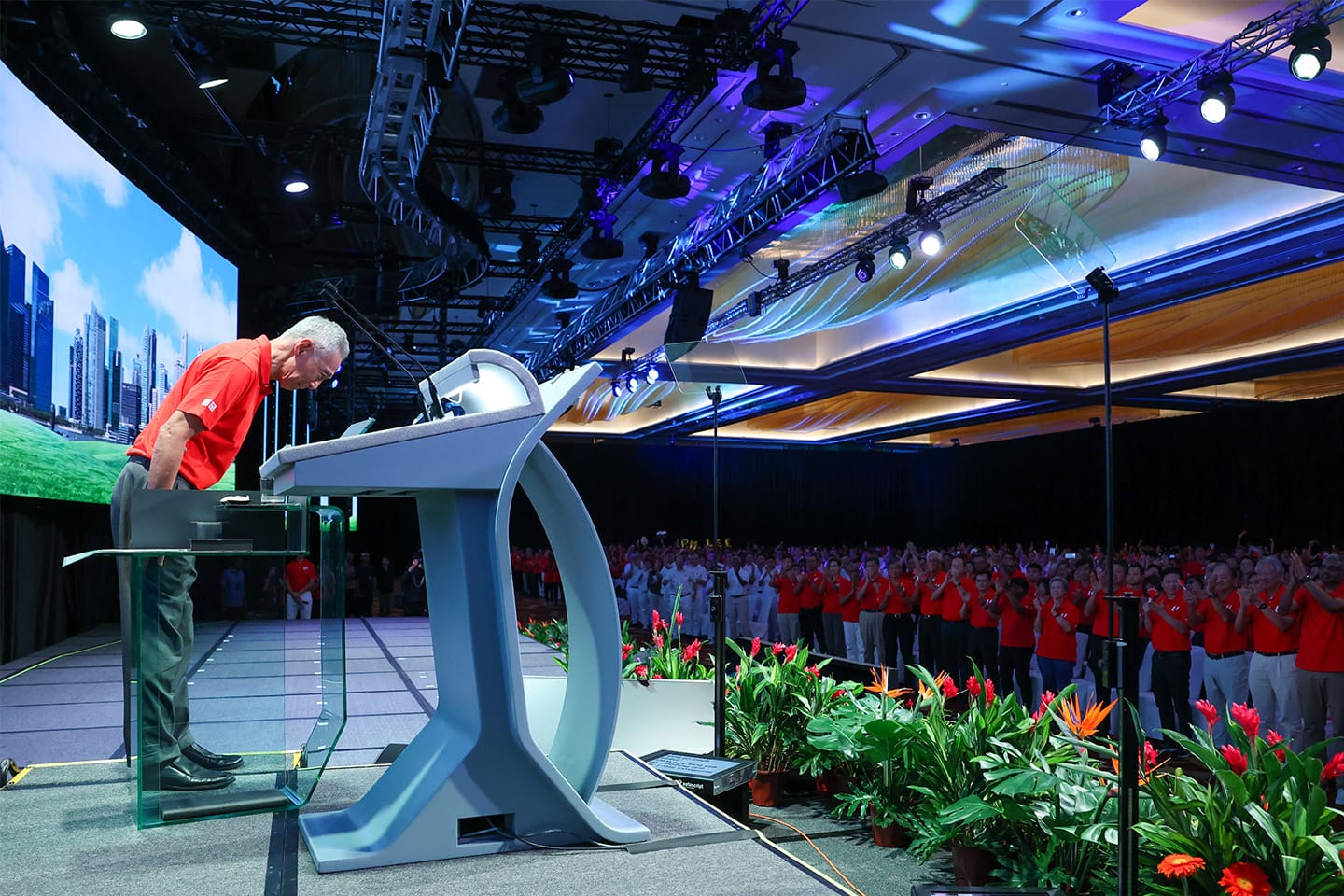 Man at podium in red shirt; cityscape screen backdrop, applauding crowd, stage plants.